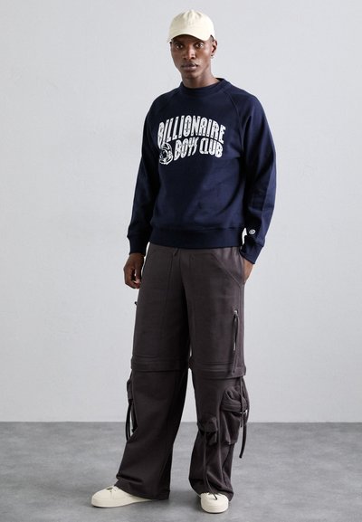 Sweat-shirt bleu marine avec texte graphique blanc ; pantalon cargo gris foncé ample avec zippers ; baskets crème ; casquette beige. Tissu texturé et détails ajustables.