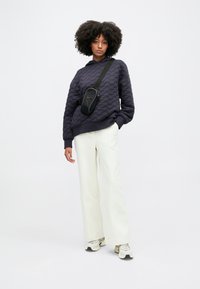 Granatowa pikowana bluza z kapturem, zestawiona z białymi spodniami o szerokich nogawkach i małą czarną torbą na ramię. Model nosi neutralne sneakersy.