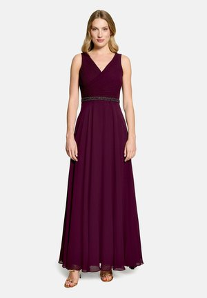 Femme portant une robe de soirée violette foncée sans manches avec un col en V, une ceinture perlée et des talons ouverts dorés, debout devant un fond uni.