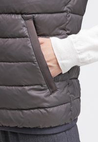 Gilet imbottito marrone scuro, con sezioni trapuntate e una tasca laterale. Il materiale appare liscio con una leggera lucentezza. La texture sembra leggera.