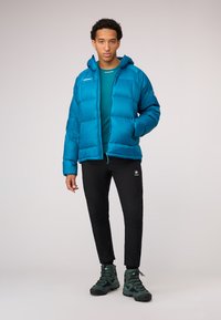 Blue puffy jacket with a hood, featuring a zip front and side pockets. Worn over a teal shirt and black pants with a logo. Dark green boots.