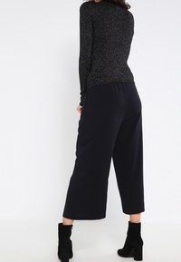 Woman wearing a black long-sleeve glittery top, wide-leg black cropped pants, and black heeled ankle boots, standing against a white background.