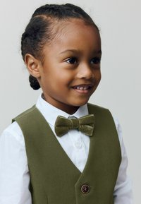 Jeune enfant souriant avec des cheveux tressés, portant une chemise blanche, un gilet vert olive et un nœud papillon en velours assorti, regardant légèrement à droite.