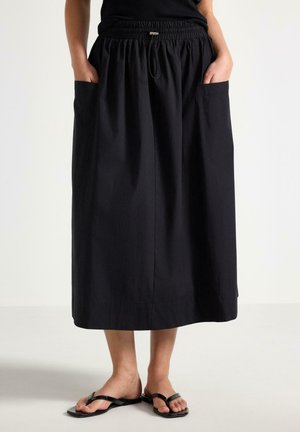 Person iført en sort mellemlang nederdel med elastisk talje og forlommer, kombineret med sorte flade thong-sandaler.