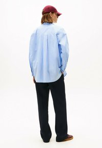 Camicia a righe blu e bianche, in stile rilassato, maniche lunghe, con dettaglio sullo scollo posteriore. Abbinata a pantaloni blu navy e un cappellino bordeaux.