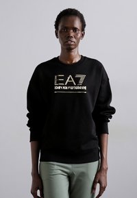 Zwarte sweatshirt met gouden Emporio Armani-logo. De stof is soepel met een relaxte pasvorm en heeft ribgebreide boorden en een zoom. Gepaarde met mintgroene broeken.