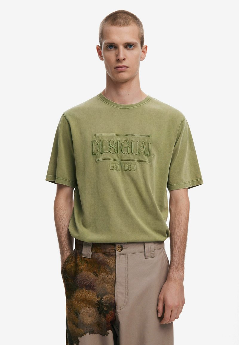 Camiseta de algodón verde oliva con bordado en relieve "DESIGUAL EST.1984" en el frente, cuello redondo y mangas de corte relajado.