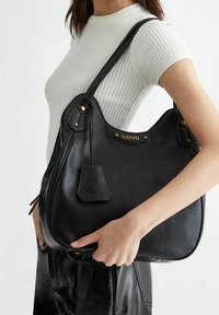 Sac à main en cuir noir avec une forme incurvée, surface texturée, accessoires en or et un accent de logo à l'avant. Bandoulière réglable pour le transport.