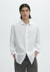 Weißes Leinenhemd mit Knopfkragen, langen Ärmeln und entspannter Passform. Kombiniert mit grauen Denim-Hosen. Minimalistisches Design.