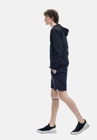 Chaqueta negra con capucha y un bolsillo frontal, combinada con pantalones cortos negros. El modelo lleva zapatillas negras y blancas, mostrando un tatuaje visible en la pierna.