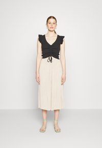 Top preto sem mangas, com babados, decote em V e detalhe de cordão, combinado com uma saia midi plissada bege e sandálias verdes de tiras.