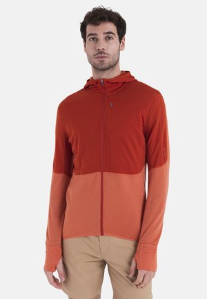 Rote Kapuzenjacke mit Reißverschluss, die ein zweifarbige Design aufweist, eine glatte Textur, lange Ärmel mit Daumenlöchern und eine Kapuze hat. Getragen über beigen Shorts.