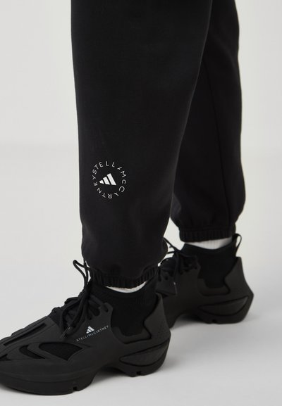Black sweatpants with elastic cuffs, featuring a white logo detail on the left leg. Paired with black athletic shoes.