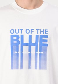 White crew neck shirt with blue text "OUT OF THE BLUE" and faded blue vertical stripes below, featuring "KARL LAGERFELD JEANS" in small white letters.