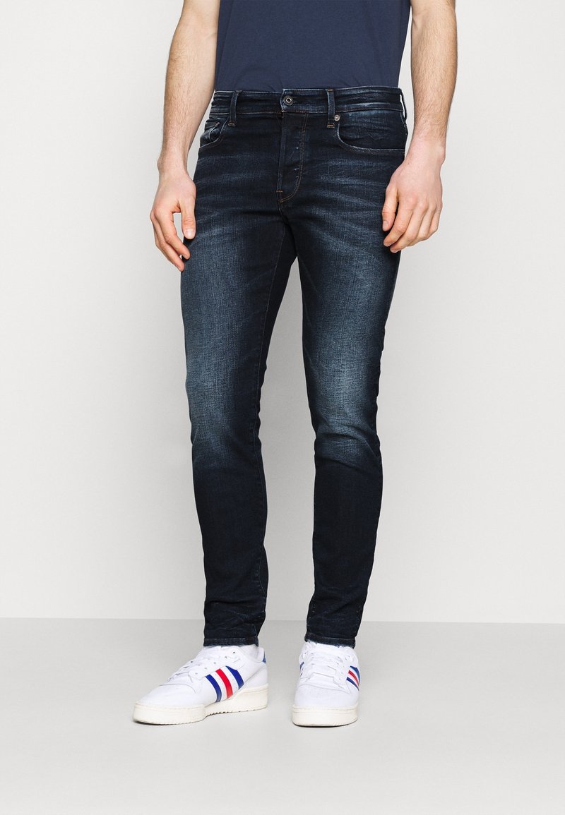 Smal passform mörkblå denimjeans med lätt blekning, femficksdesign och bältesöglor, tillsammans med vita sneakers med röda och blå ränder.