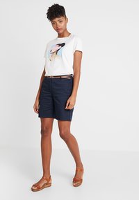 T-shirt blanc avec un imprimé graphique coloré, associé à un short bleu marine et des sandales compensées marron ; comprend une fine ceinture marron.