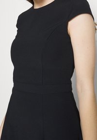 Robe noire ajustée avec manches courtes, corsage structuré et taille bien définie. Texture lisse et design minimaliste sans ornements.