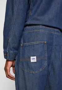 Jean en denim bleu foncé, avec des coutures contrastantes orange, des poches arrière aux bords arrondis, et une étiquette rectangulaire de la marque Lee.