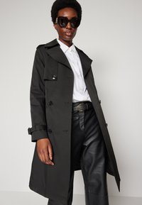 Schwarzer Trenchcoat mit Knopfverschluss und Gürtel, ausgestattet mit einem breiten Kragen und langen Ärmeln. Kombiniert mit einem weißen Hemd und schwarzen Lederhosen.