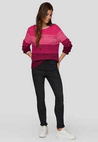 Strickpullover mit einem Farbverlauf in verschiedenen Rosatönen und gerippten Bündchen, kombiniert mit schwarzen Skinny-Jeans und weißen Sneakers, steht vor einem grauen Hintergrund.