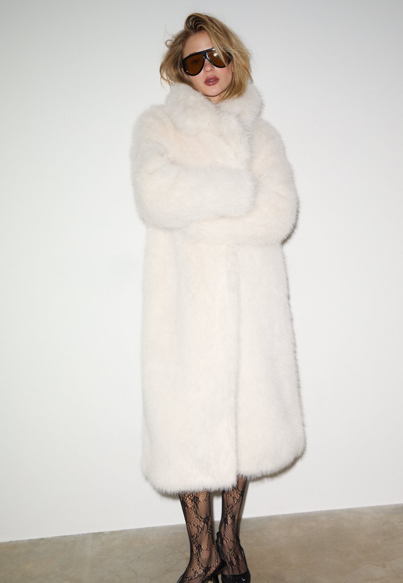 Cappotto in pelliccia sintetica bianca con collo alto, caratterizzato da una texture morbida e soffice. Calze a rete nere e scarpe nere eleganti completano il look.