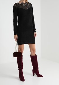Robe-pull noire avec un décolleté en dentelle, des manches longues et une coupe ajustée. Le mannequin porte des bottes longues bordeaux à la coupe décontractée et tient un petit sac en velours.