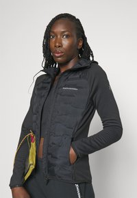 Sort hybridjakke med en puffy front, tætsiddende ærmer, lynlåslommer og hætte. Logo på brystet, struktureret stof og kontrasterende materialer.