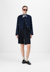 Jachetă croppată din lână bleumarin, cu nasturi aurii, asociată cu pantaloni scurți negri ajustați. Modelul poartă un fular cu model și ochelari de soare, cu mocasini negri.