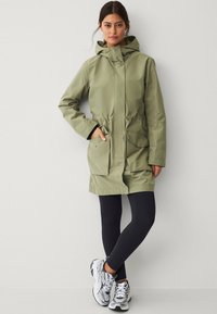 Olijfgroene waterdichte parka met capuchon, koord in de taille en twee voorkeepockets, gecombineerd met zwarte leggings en gestreepte sportschoenen.