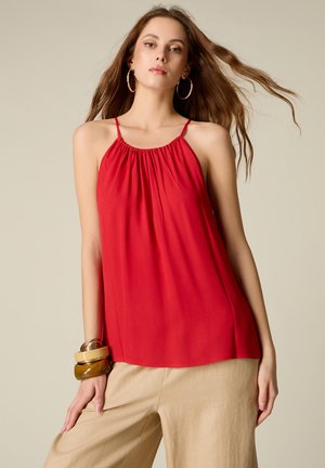 Kvinde med langt brunt hår iført en rød ærmeløs haltertop, beige bukser, store ringøreringe og stablede brune armbånd på venstre håndled.