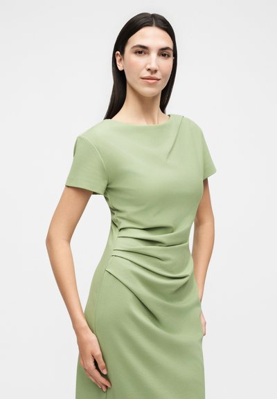 Γυναίκα με μακριά σκούρα μαλλιά που φοράει εφαρμοστό ανοιχτό πράσινο φόρεμα με κοντά μανίκια, στέκεται με το ένα χέρι στη μέση της μπροστά σε λευκό φόντο.