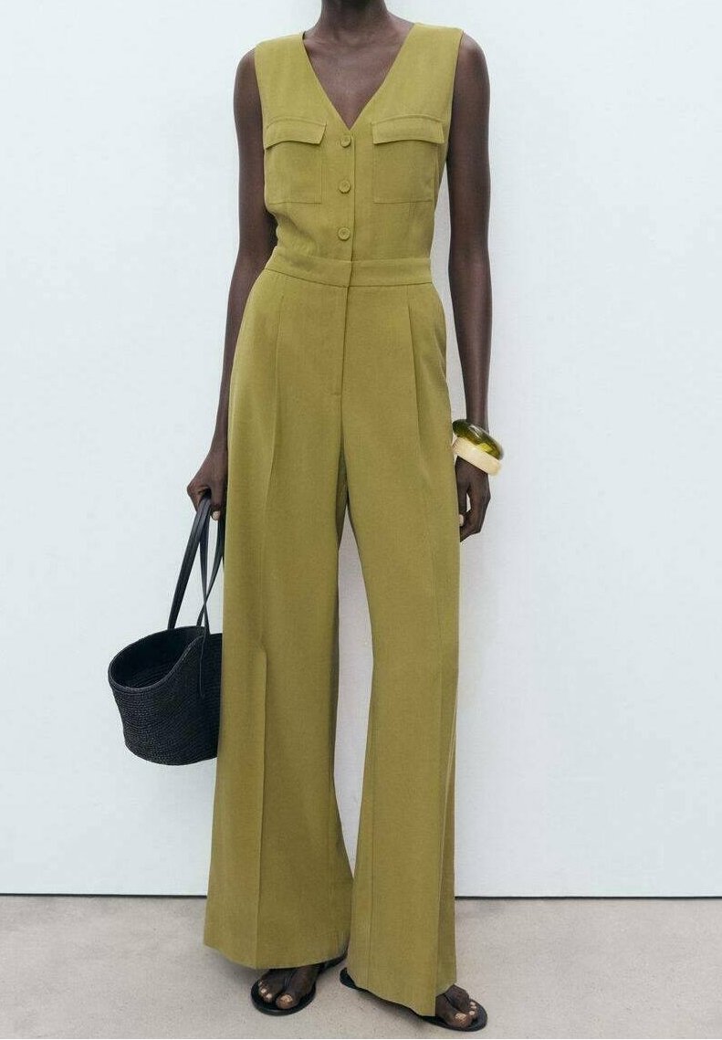 Femme portant une combinaison sans manches vert olive à jambes larges, tenant un sac à main noir et portant des sandales et bracelets noirs.