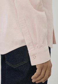 Chemise en velours côtelé rose clair avec un poignet boutonné et un côtelé vertical. La chemise est associée à un jean en denim foncé.
