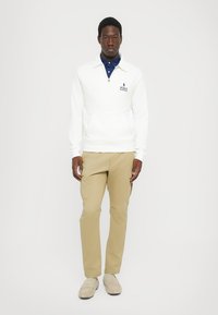 Valge kvart-zipp pullover koos logoga, kantud tumesinise kraega särke. Kombineeritud beežide pükstega ja heleda jalanõudega. Lihtne disain ja lõige.