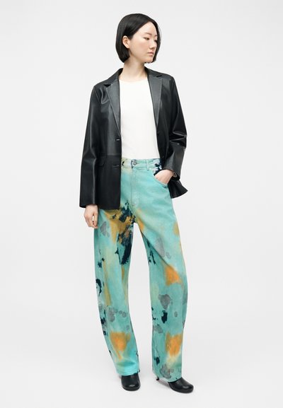 Γυναίκα με κοντά μαύρα μαλλιά, φοράει μαύρο δερμάτινο σακάκι, λευκό τοπ, πολύχρωμο παντελόνι tie-dye σε μπλε και πορτοκαλί με φαρδιά μπατζάκια, και μαύρα μπότες με τακούνι.