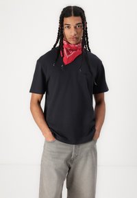 Tricou negru cu mânecă scurtă și decolteu rotund, fabricat din bumbac. Modelul poartă o bandană roșie la gât și blugi gri, largi.
