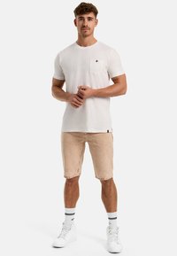 Hvid bomulds t-shirt med rund hals og lomme, parret med lysebeige denimshorts. Modellen bærer hvide sneakers med sorte striber på sokkerne.