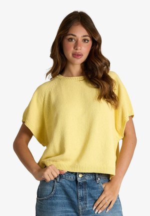 Giovane donna con lunghi capelli castani indossa un maglione giallo largo a maniche corte e jeans blu con la mano sul fianco.