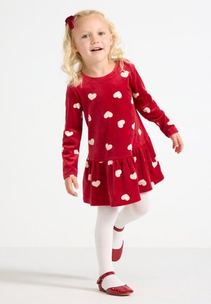 Rotes Samtkleid mit weißen Herzmustern, langen Ärmeln und gewelltem Saum. Kombiniert mit weißen Strumpfhosen und roten Schuhen. Kind in einer verspielten Pose.