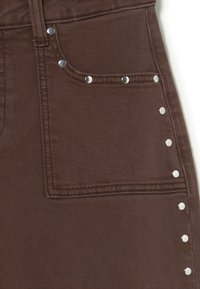 Jeans de mezclilla marrones con un bolsillo frontal, que presentan acentos de tachuelas plateadas a lo largo de las costuras laterales y una tela suave y texturizada.