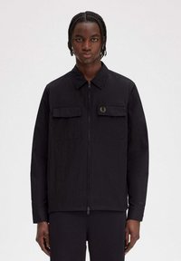 Giacca nera leggera con zip frontale, colletto e due tasche anteriori. Presenta una discreta patch con logo sulla parte sinistra del petto.