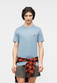 Νεαρό άτομο που φοράει ανοιχτό μπλε μπλουζάκι με μικρό λογότυπο ελάφι, γκρι σορτς και κόκκινο-μαύρο καρό πουκάμισο δεμένο γύρω από τη μέση.