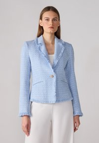 Lyseblå tweed-blazer med frynsete revers, to frontlommer og en dekorativ gullknapp, kombinert med hvite bukser.