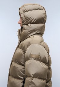 Giacca puffer imbottita beige con collo alto e cappuccio. Texture liscia e lucida, con un patch con logo sulla manica superiore e dettagli con cordoncino.