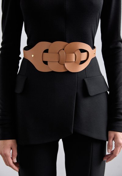 Curea lată din piele bej cu un design în bucle interconectate, dotată cu accesorii aurii, purtată peste o ținută neagră texturată cu accente de buzunare.