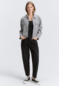 Mujer vestida con una chaqueta gris de botones, una camiseta sin mangas negra, pantalones negros de tiro alto con un lazo, y zapatillas negras de corte bajo con suelas blancas.