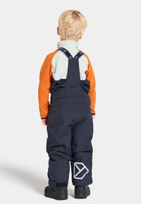 Mörkblå snöoverall med justerbara remmar och bakfickor, i kombination med en lager-på-lager-topp i orange och mintgrön. Svarta stövlar.