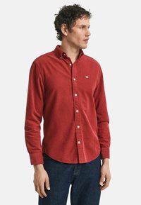 Homme aux cheveux bouclés portant une chemise en velours côtelé rouge à bouton et un jean bleu marine, regardant vers la droite contre un fond blanc uni.