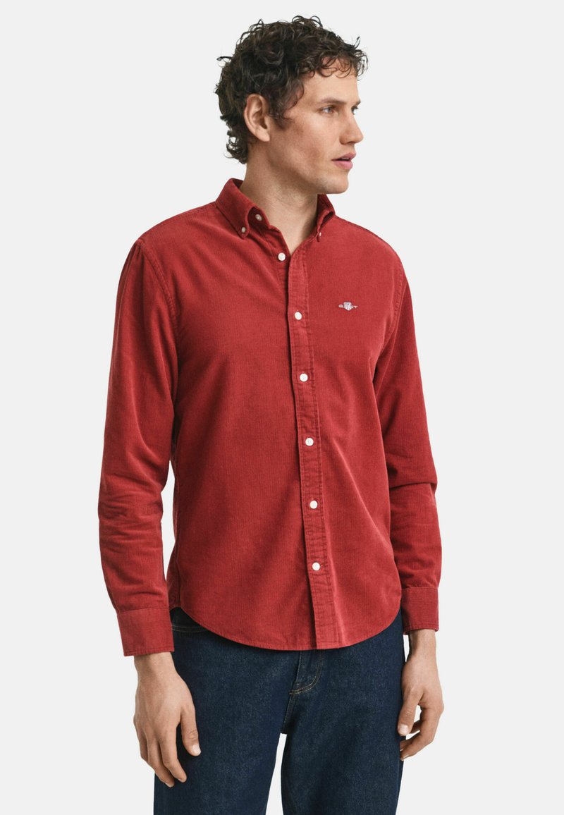 Homme aux cheveux bouclés portant une chemise en velours côtelé rouge à bouton et un jean bleu marine, regardant vers la droite contre un fond blanc uni.