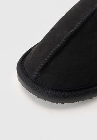 Svart tygtoffel med rund tå, sydda kanter och texturerad gummisula, som framhäver en enkel och funktionell design.